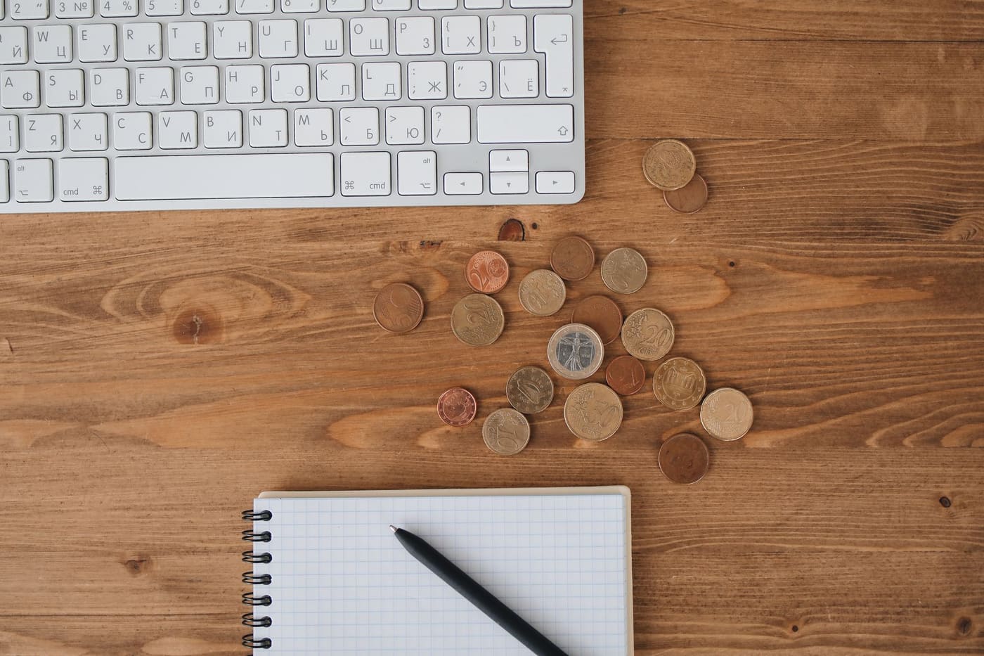 Coins scattered on a wooden desk beside a keyboard and a blank notebook