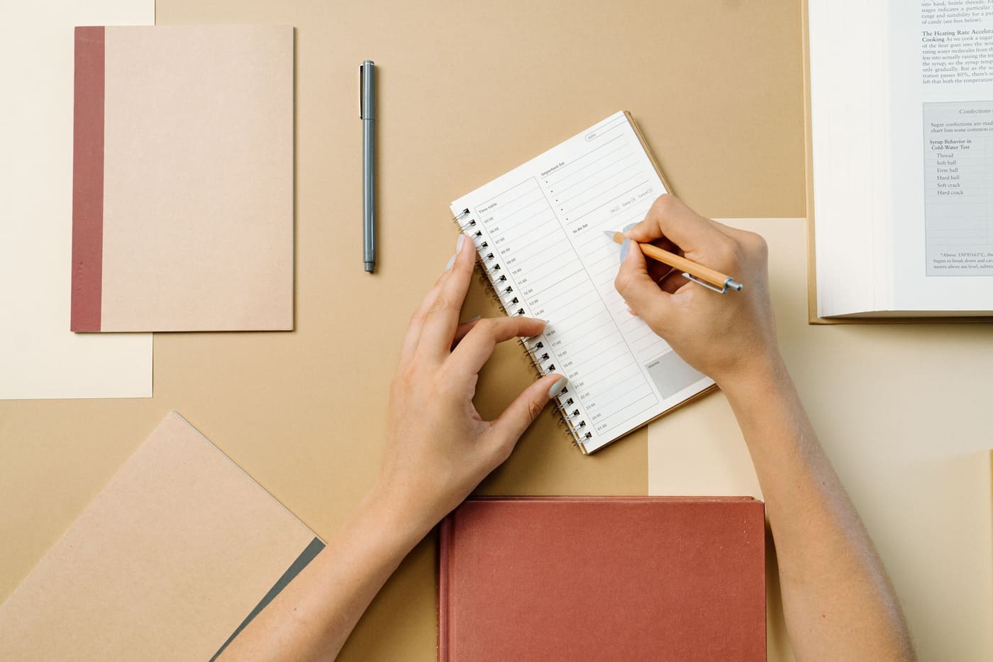 Hands writing in a spiral notebook on a desk with other notebooks