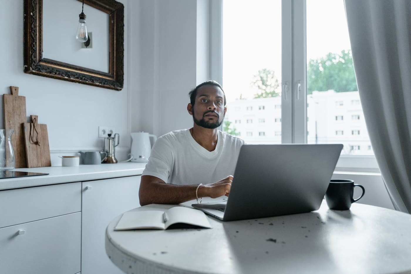 A man working on a laptop at a white kitchen table beside a window