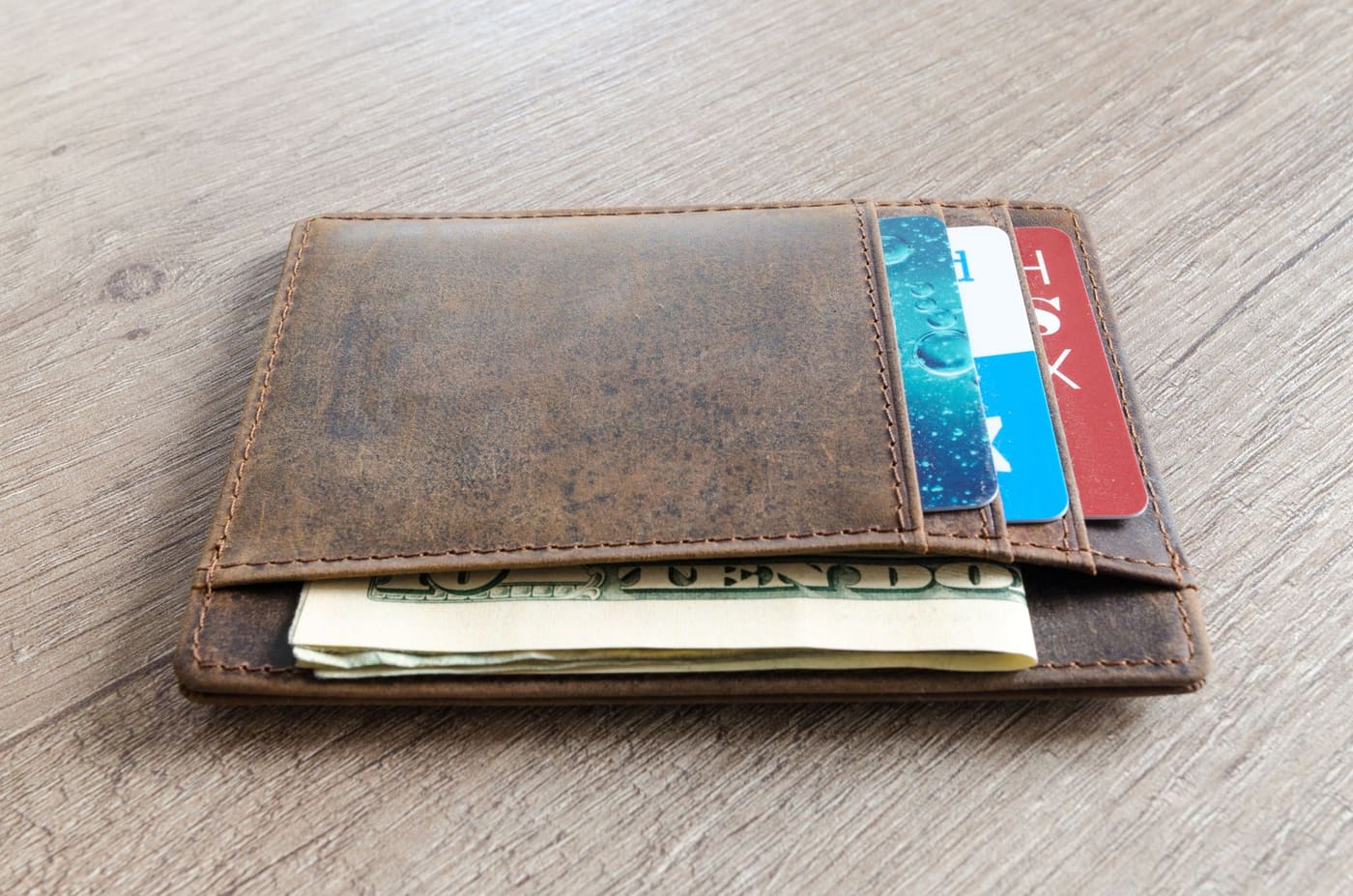 A worn brown leather wallet holding cash and cards on a wooden surface