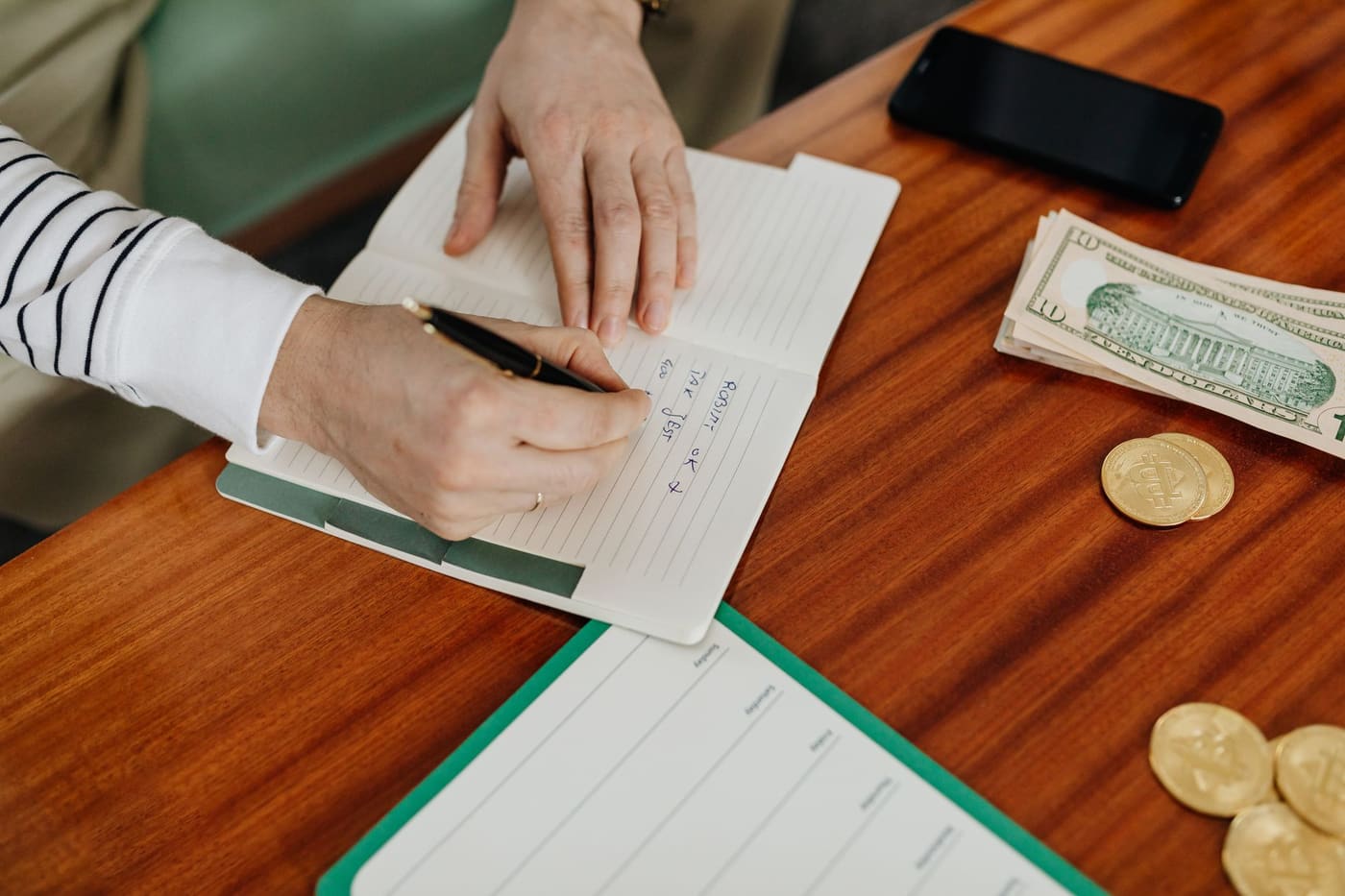 A person writing in a notebook on a wooden table beside cash, coins and a phone