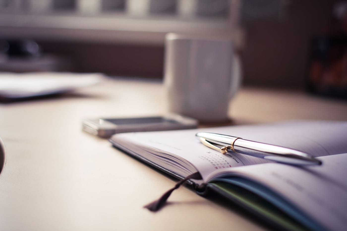 An open planner with a pen and a coffee mug on a desk