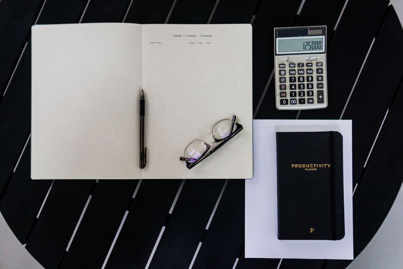 A flat lay of an open notebook, a calculator, a planner and reading glasses on a dark table